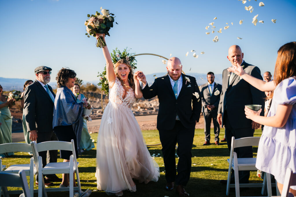 Newlyweds walking down the aisle with happiness, carrying bouquets of flowers, and petals being joyfully tossed in the air at Revere Golf Club