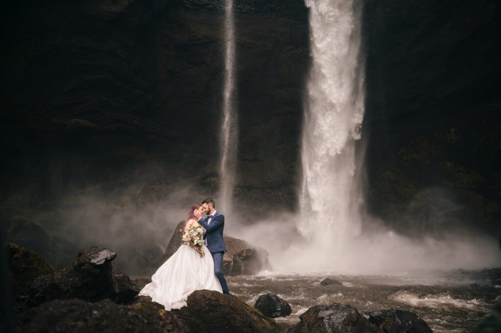 In the heart of Iceland, a couple stands face to face, their love shining brightly. Behind them, a majestic waterfall adds a touch of enchantment to their intimate elopement moment.